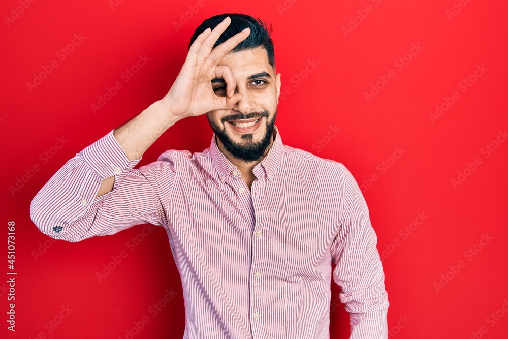 Handsome man with beard wearing casual red shirt smiling happy doing ok sign with hand on eye looking through fingers Handsome man with beard wearing casual red shirt smiling happy doing ok sign with hand on eye looking through fingers