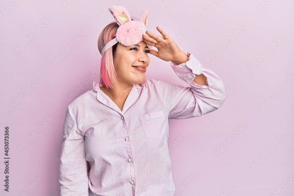Hispanic woman with pink hair wearing sleep mask and pajama very happy and smiling looking far away with hand over head. searching concept. Hispanic woman with pink hair wearing sleep mask and pajama very happy and smiling looking far away with hand over head. searching concept.
