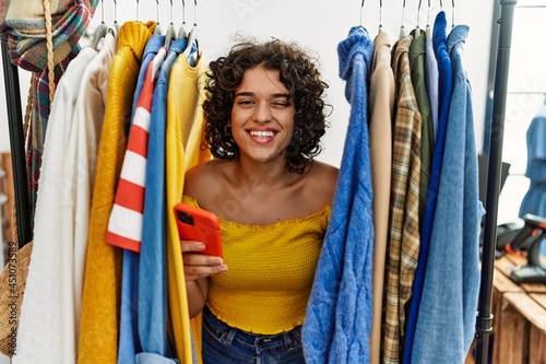Young hispanic woman searching clothes on clothing rack using smartphone winking looking at the camera with sexy expression, cheerful and happy face.