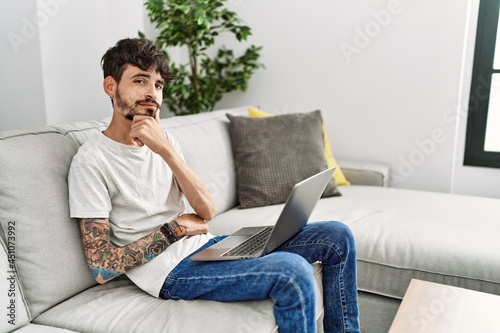 Hispanic man with beard sitting on the sofa looking confident at the camera with smile with crossed arms and hand raised on chin. thinking positive.