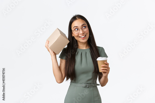 Intrigued smiling asian female drinking coffee, holding mug and shaking box as trying guess what inside, looking curious, receiving parcel from online store, standing white background photo