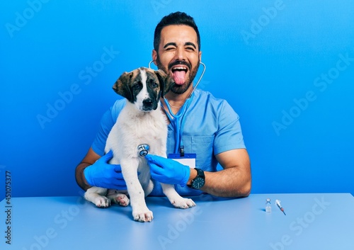 Fototapeta Naklejka Na Ścianę i Meble -  Handsome hispanic veterinary man with beard checking dog health using stethoscope sticking tongue out happy with funny expression.