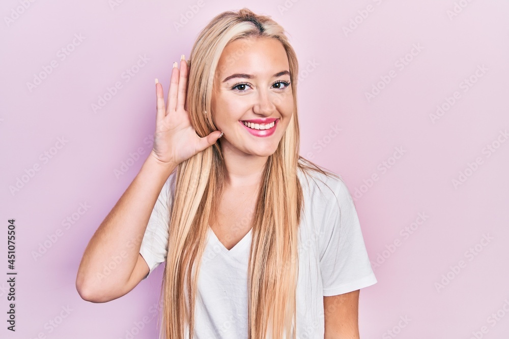Young blonde girl wearing casual white t shirt smiling with hand over ear listening and hearing to rumor or gossip. deafness concept. Young blonde girl wearing casual white t shirt smiling with hand over ear listening and hearing to rumor or gossip. deafness concept.
