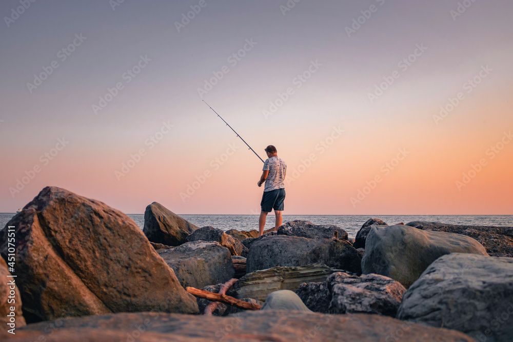 A fisherman catches fish at sunset. Active recreation in nature. A man with a fishing rod is fishing on the rocks near the sea.
