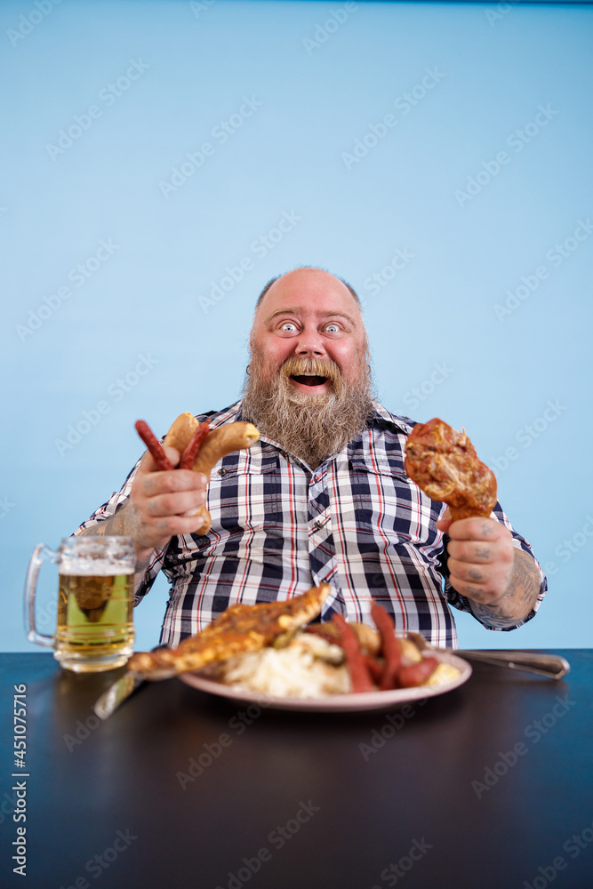 Excited obese man holds chicken leg and bunch of sausages sitting at table with greasy food and beer on light blue background in studio Excited obese man holds chicken leg and bunch of sausages sitting at table with greasy food and beer on light blue background in studio