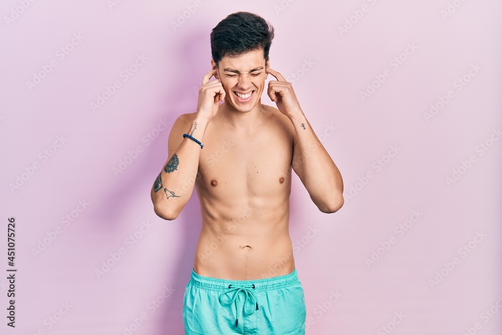 Fototapeta premium Young hispanic man wearing swimwear shirtless covering ears with fingers with annoyed expression for the noise of loud music. deaf concept.