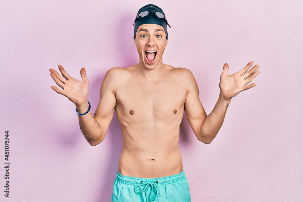 Young hispanic man wearing swimwear and swimmer glasses celebrating crazy and amazed for success with arms raised and open eyes screaming excited. winner concept