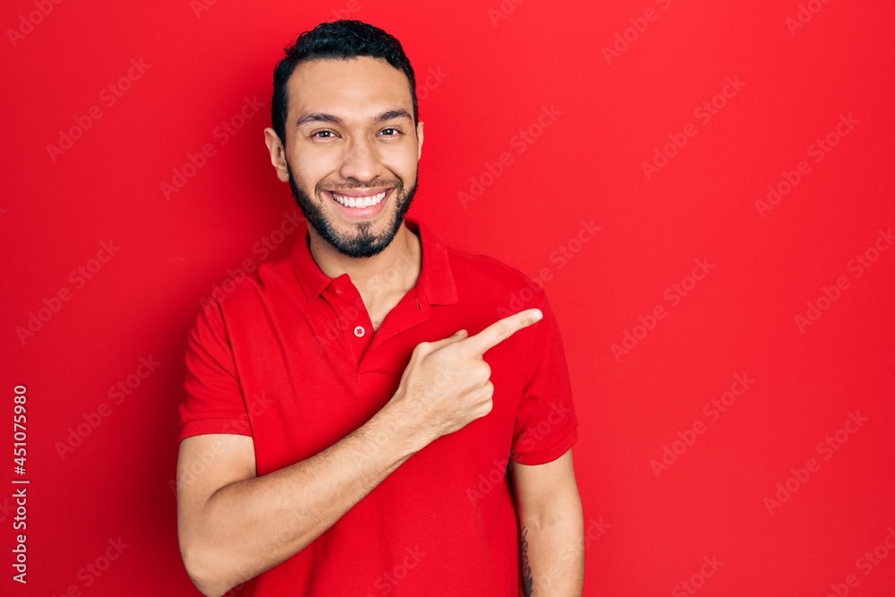 Hispanic man with beard wearing casual red t shirt cheerful with a smile of face pointing with hand and finger up to the side with happy and natural expression on face Hispanic man with beard wearing casual red t shirt cheerful with a smile of face pointing with hand and finger up to the side with happy and natural expression on face
