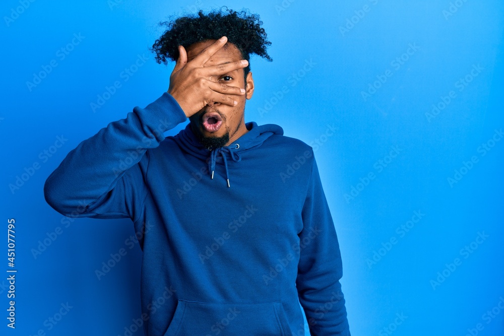 Young african american man with beard wearing casual sweatshirt peeking in shock covering face and eyes with hand, looking through fingers afraid Young african american man with beard wearing casual sweatshirt peeking in shock covering face and eyes with hand, looking through fingers afraid