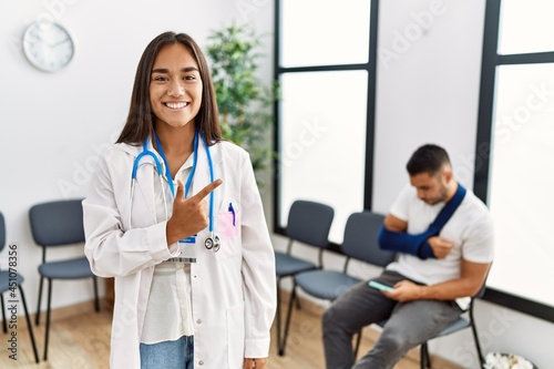 Young asian doctor woman at waiting room with a man with a broken arm cheerful with a smile of face pointing with hand and finger up to the side with happy and natural expression on face