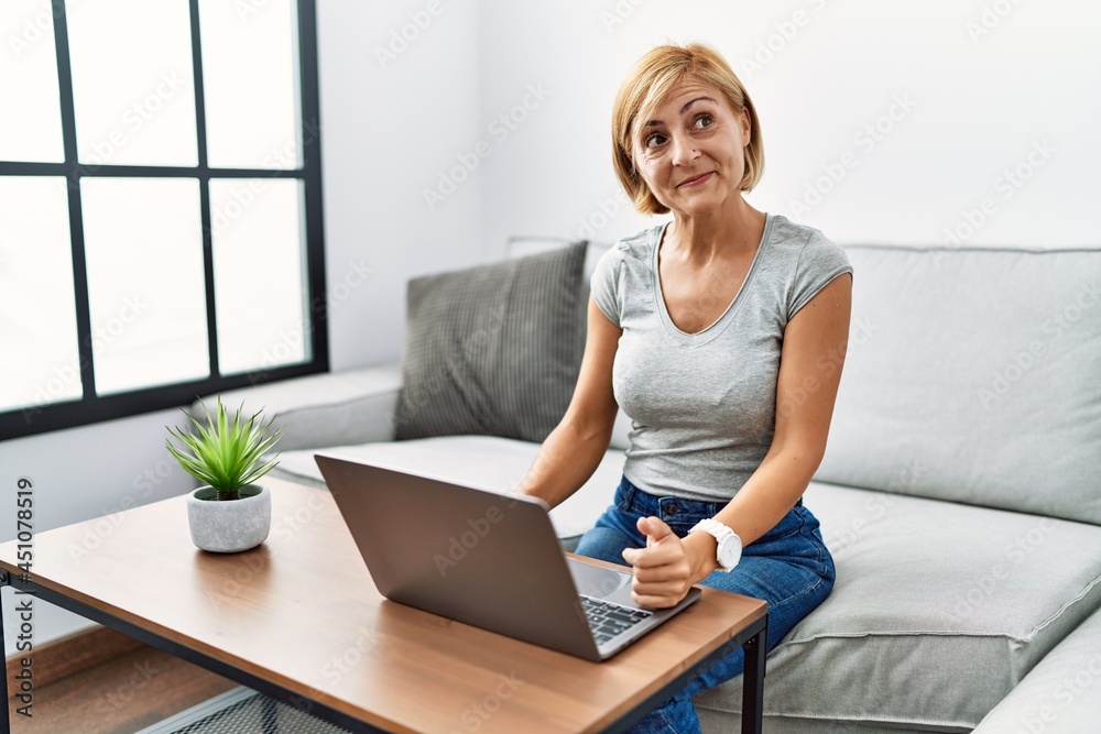 Middle age blonde woman using laptop at home smiling looking to the side and staring away thinking. Middle age blonde woman using laptop at home smiling looking to the side and staring away thinking.