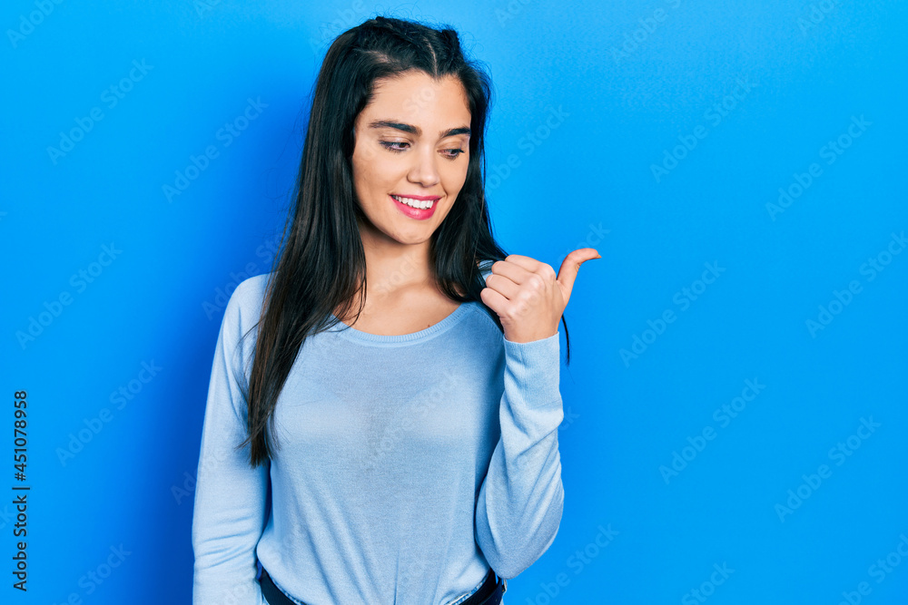 Young hispanic girl wearing casual clothes smiling with happy face looking and pointing to the side with thumb up. Young hispanic girl wearing casual clothes smiling with happy face looking and pointing to the side with thumb up.