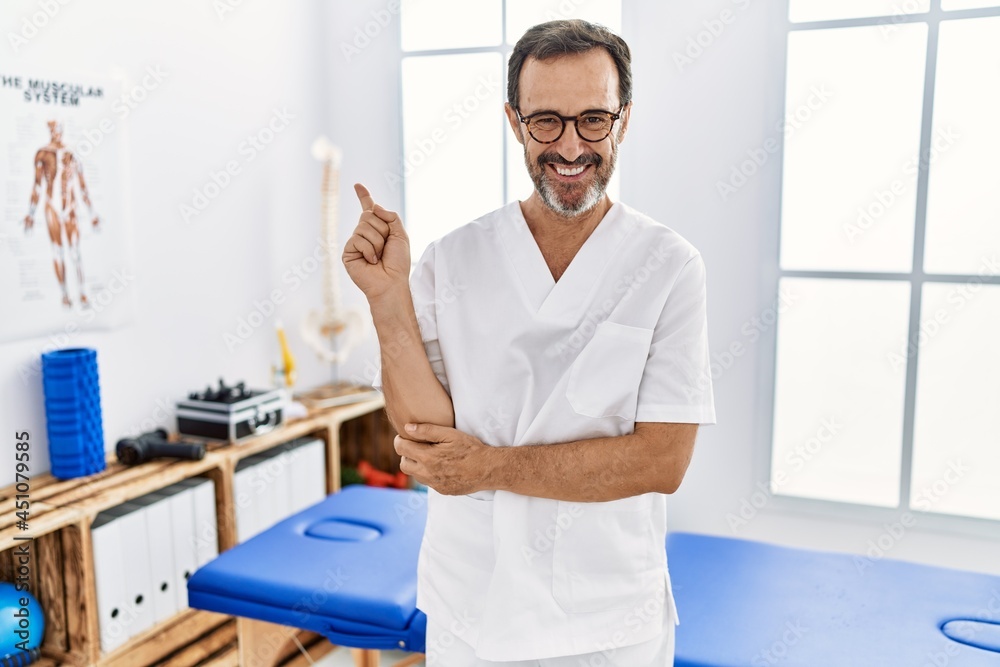 Middle age man with beard working at pain recovery clinic with a big smile on face, pointing with hand and finger to the side looking at the camera. Middle age man with beard working at pain recovery clinic with a big smile on face, pointing with hand and finger to the side looking at the camera.