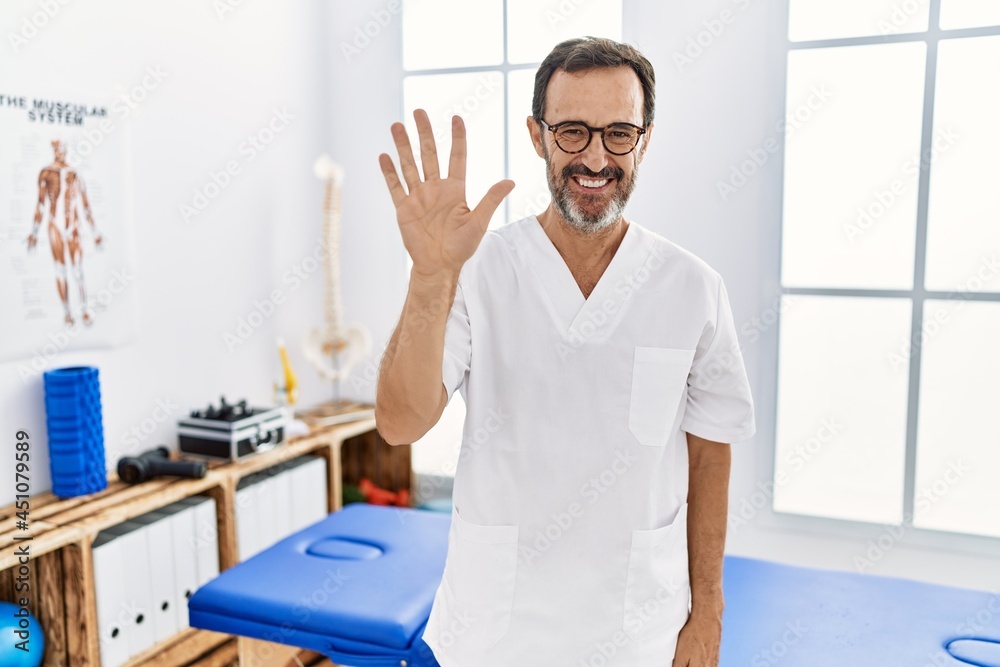 Middle age man with beard working at pain recovery clinic showing and pointing up with fingers number five while smiling confident and happy. Middle age man with beard working at pain recovery clinic showing and pointing up with fingers number five while smiling confident and happy.