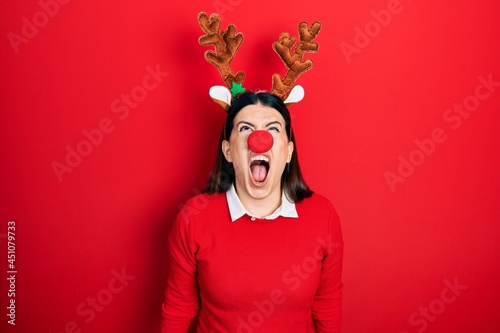 Young hispanic woman wearing deer christmas hat and red nose angry and mad screaming frustrated and furious, shouting with anger. rage and aggressive concept.