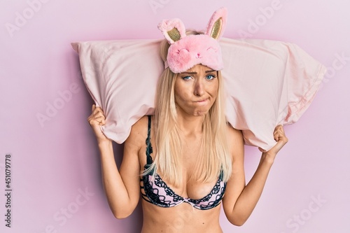 Young blonde woman wearing lingerie and sleep mask holding pillow skeptic and nervous, frowning upset because of problem. negative person.