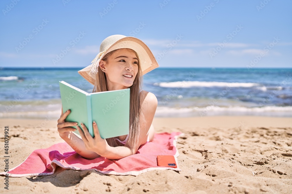 Young chinese girl reading book lying on the towel at the beach.
