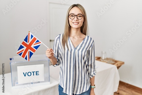 Asian young woman at political campaign election holding uk flag looking positive and happy standing and smiling with a confident smile showing teeth