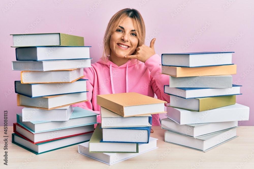Young caucasian woman sitting on the table with books smiling doing phone gesture with hand and fingers like talking on the telephone. communicating concepts. Young caucasian woman sitting on the table with books smiling doing phone gesture with hand and fingers like talking on the telephone. communicating concepts.