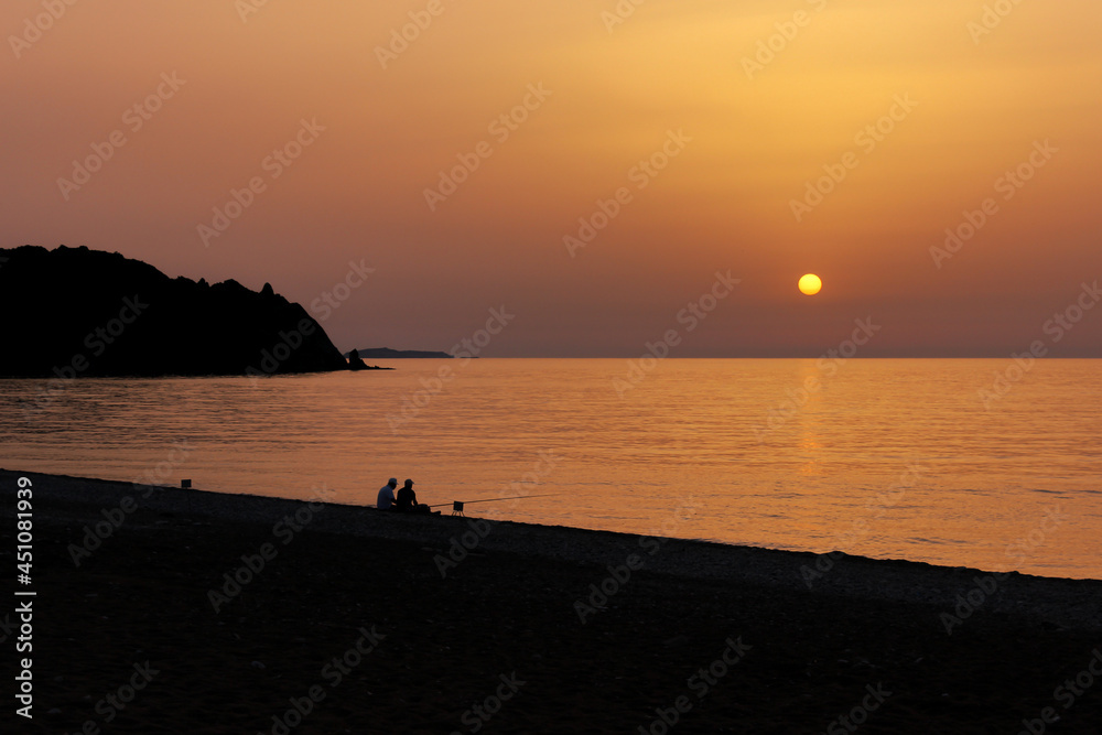 Silhouette of two men fishing on the beach during the sunrise. Sun rises over the Mediterranean Sea on a lovely summer morning. Selective focus.
