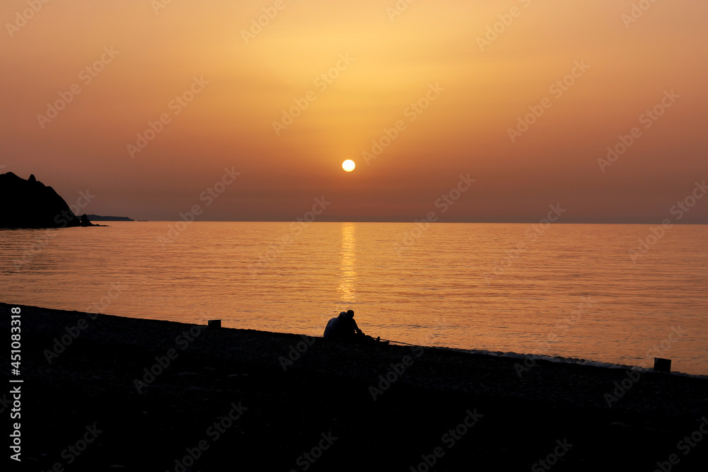 Silhouette of two people sitting on the beach during the sunrise. Sun rises over the Mediterranean Sea on a lovely summer morning. Selective focus.