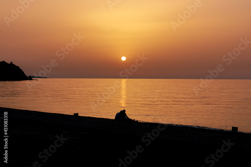Silhouette of two people sitting on the beach during the sunrise. Sun rises over the Mediterranean Sea on a lovely summer morning. Selective focus.