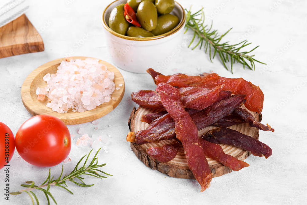 Jerky meat strips with spices, green olives, rosemary on wooden plate on a grey stone background. Tipical Italian food coppiette is a spiced pork meat strips. Snacks for beer, close up Jerky meat strips with spices, green olives, rosemary on wooden plate on a grey stone background. Tipical Italian food coppiette is a spiced pork meat strips. Snacks for beer, close up