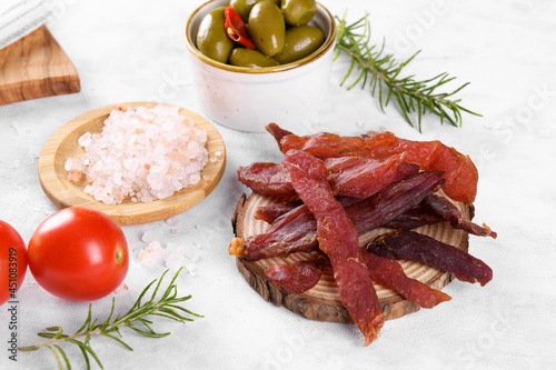 Jerky meat strips with spices, green olives, rosemary on wooden plate on a grey stone background. Tipical Italian food coppiette is a spiced pork meat strips. Snacks for beer, close up photo