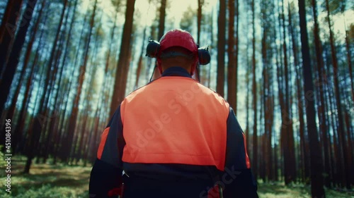 A forest worker in overalls, a helmet and a vest with his back to the camera walks through the forest. Live action camera. The lumberjack goes to his workplace