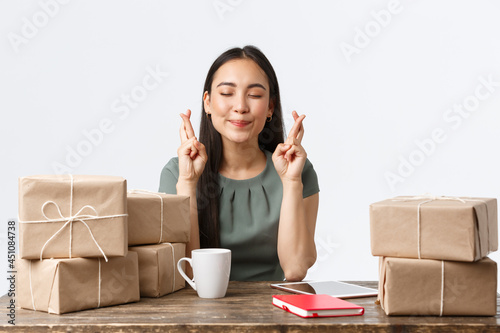 Small business owners, startup and e-commerce concept. Hopeful asian businesswoman praying for her online store be popular, sitting with packed orders for shipping, hold fingers crossed photo