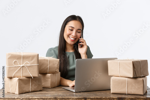 Small business owners, startup and e-commerce concept. Smiling asian female store manager, entrepreneur with online shop answering client calls, managing orders via laptop, packing items for shipping photo