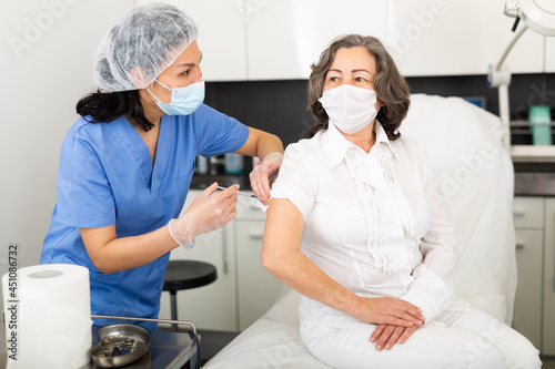 Professional nurse in blue uniform and protective face mask giving antiviral injection to aged woman in medical office. Vaccination  immunization and disease prevention concept