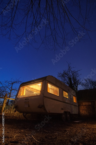 Old white caravan trailer with yellow illumination in backyard at stary blue night