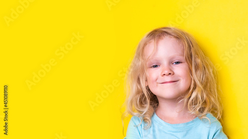 portrait beautiful curly funny little girl with blond curly hair wearing blue t-shirt on yellow background with copy space