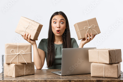 Small business owners, startup and e-commerce concept. Amazed young asian woman open online store, sitting table with laptop, managing clients orders, packing items in boxes for shipping photo