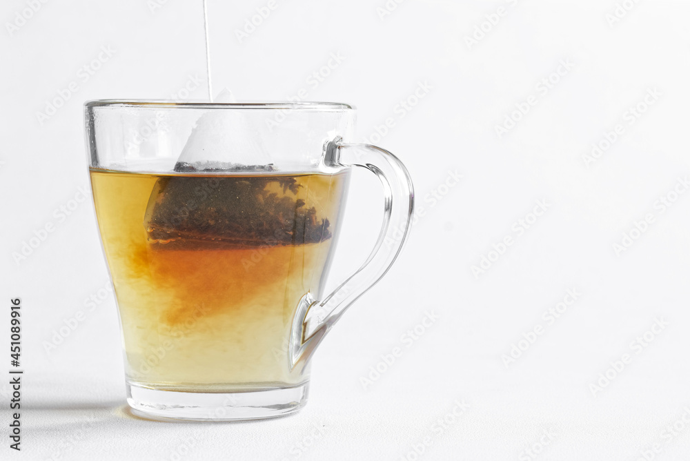 Tea bag - a pyramid of black tea is brewed in hot water in a transparent mug. White background. Free space for an inscription Tea bag - a pyramid of black tea is brewed in hot water in a transparent mug. White background. Free space for an inscription
