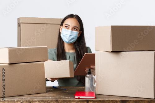 Small business owners, e-commerce and covid-19 preventing virus concept. Smiling asian businesswoman with store, packing orders in medical mask and gloves, using hand sanitizer before touching parcel photo