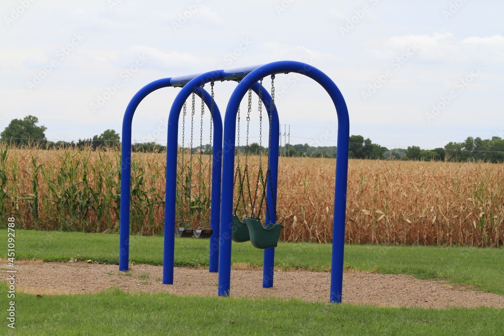 Fototapeta premium playground in the park with swings on blue poles with a corn field in the background. In Sterling Kansas USA.