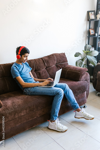 Hispanic teenager boy with headphones sitting on sofa while using laptop at home in Latin America © Marcos