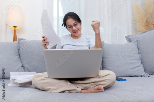 Asian woman celebrating online win success looking laptop at home. Work from home business women sitting on sofa in living room working with laptop online. Young women excited and glad of success 