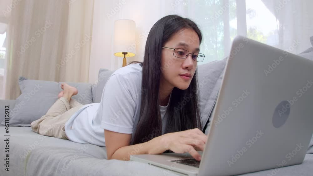 Asian woman relaxing at home on the couch and using laptop computer. Smiling woman using laptop on sofa at home. Women working at home with computer at living room Asian woman relaxing at home on the couch and using laptop computer. Smiling woman using laptop on sofa at home. Women working at home with computer at living room