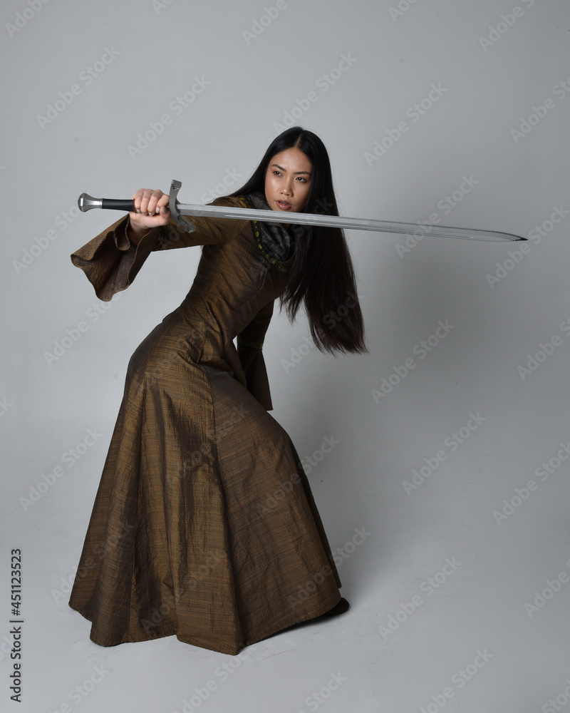 Full length portrait of beautiful young asian woman with long hair wearing medieval fantasy gown costume. Graceful standing posing holding a sword, isolated on studio background. Full length portrait of beautiful young asian woman with long hair wearing medieval fantasy gown costume. Graceful standing posing holding a sword, isolated on studio background.