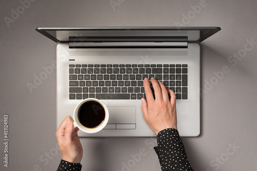 First person top view photo of woman's hands typing on laptop keyboard and holding cup of coffee on isolated grey background