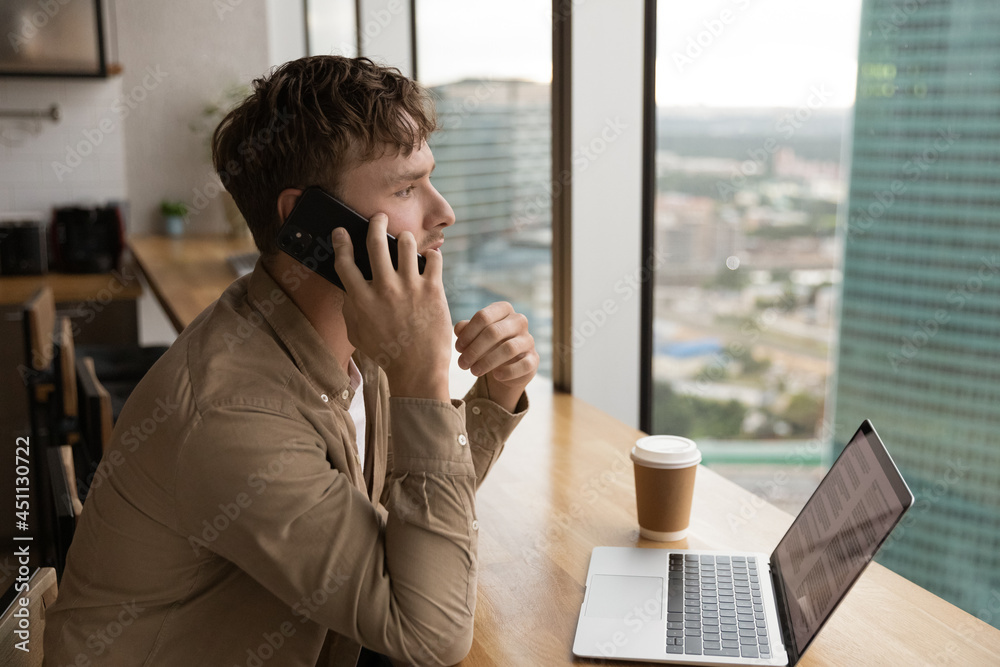 Side view millennial man sit in modern office room holding wireless device talking to client having serious conversation on smart phone, working, making business remote talk use modern tech concept Side view millennial man sit in modern office room holding wireless device talking to client having serious conversation on smart phone, working, making business remote talk use modern tech concept
