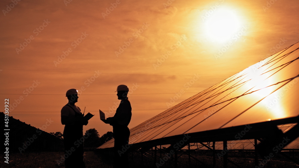 Two unidentified solar power engineers talking to each other at sunset. Couple of workers examining solar batteries and communicating on business. Ecological future. Sustainable energy.
