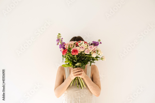 Colourful flowers bouquet in female hands on white background. Young woman in neutral beige washed linen dress hide face. Aesthetic beauty and fashion floral concept. Face hidden by flower bouquet
