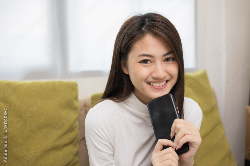 Happy young asian woman looking at screen of smartphone sitting on sofa in living room she shopping online. She very happy. Teenage girl reading news from mobile phone feeling excited. Happy young asian woman looking at screen of smartphone sitting on sofa in living room she shopping online. She very happy. Teenage girl reading news from mobile phone feeling excited.