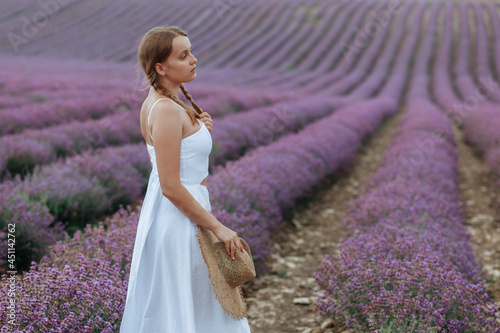 Fototapeta Naklejka Na Ścianę i Meble -  A beautiful young woman in a white dress with a hat walks through a lavender field