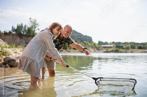 Happy preteen girl and grandfather with fishing net on summer holiday by lake.