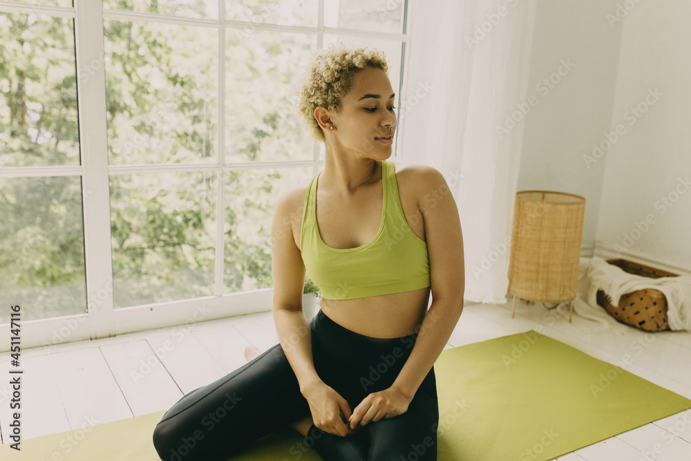 Picture in profile of trendy cute African fitness trainer doing gymnastics at home panoramic window on background, doing stretching on green mat in sport top and leggings. Healthy lifestyle. Wellness Picture in profile of trendy cute African fitness trainer doing gymnastics at home panoramic window on background, doing stretching on green mat in sport top and leggings. Healthy lifestyle. Wellness